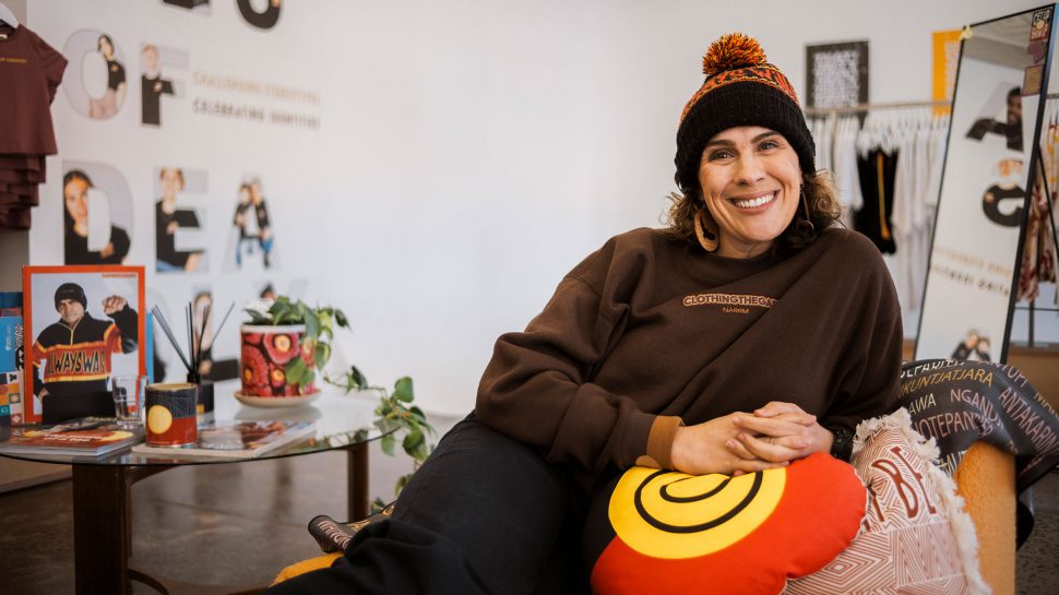 Clothing the Gaps co-founder, Laura Thompson, smiles as she lounges on a chair at work adorned in Clothing the Gaps clothes and merchandise.