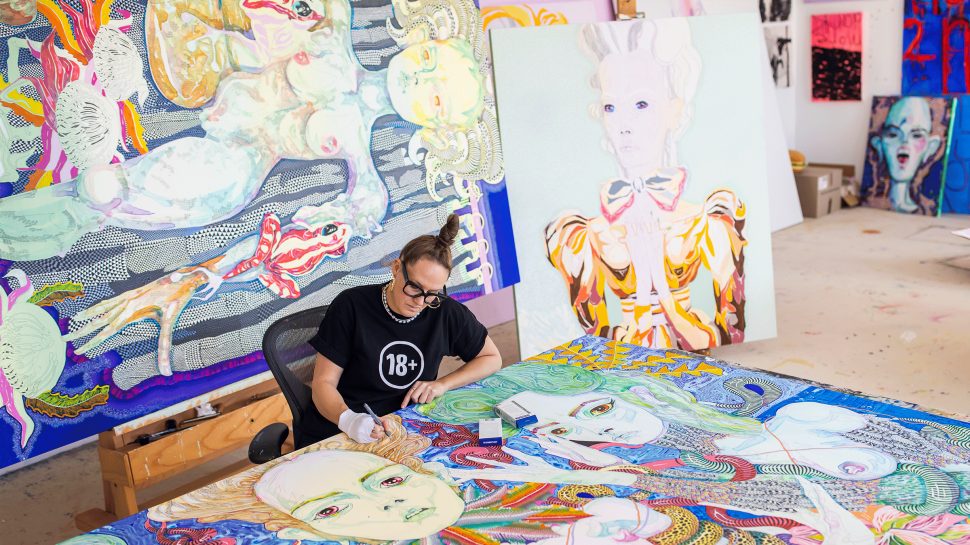 A woman wearing a black T-shirt is working at a painting in on a large desk. She’s surrounded by canvases of other paintings.