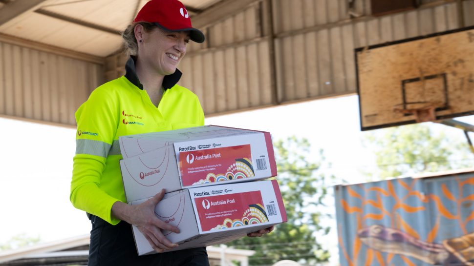 A female Australia Post team member wearing high-vis and a red cap carries boxes. Boxes feature Australia Post label with Indigenous artwork.