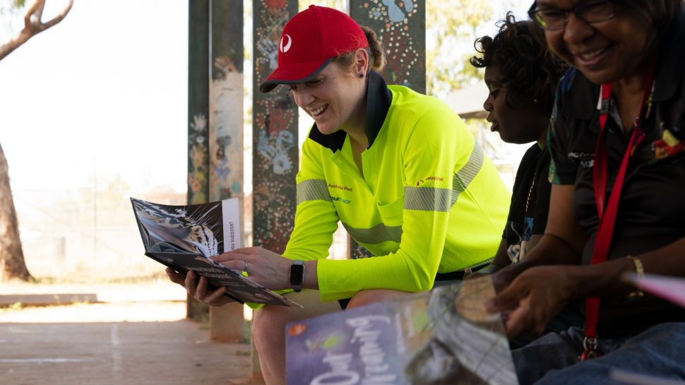 Australia Post team member wearing high-vis and a red cap sits with Indigenous community members as they flip through books.   