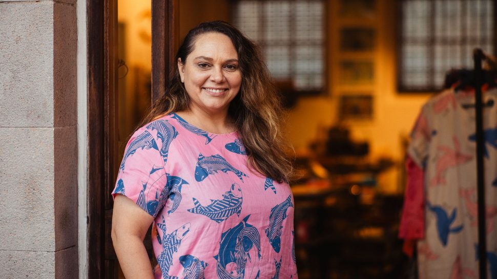 Magpie Goose co-owner, Amanda Hayman, smiles in the doorway of her business while wearing a Magpie Goose dress adorned with Aboriginal artwork.