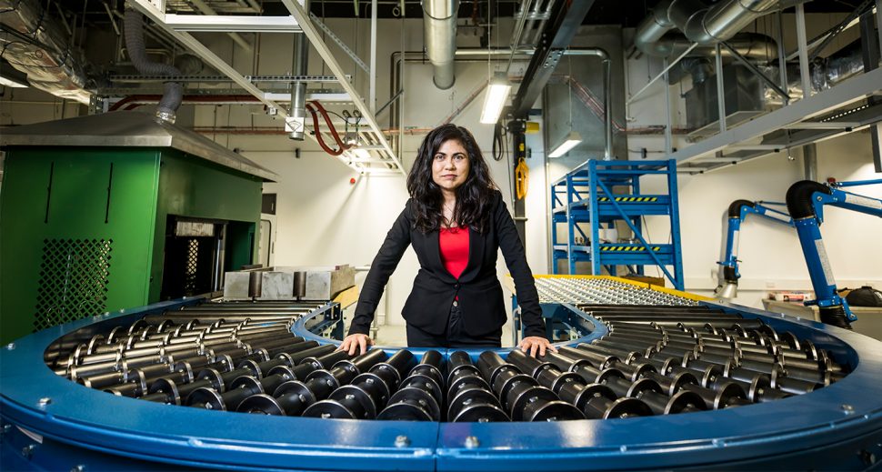 An olive-skinned woman in a red top and black blazer standing in a lab.