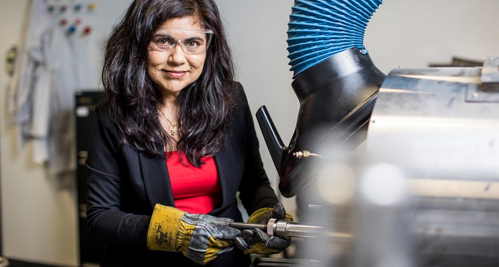 An olive-skinned woman in a red top, black blazer protective eyewear and yellow gloves working on a machine.