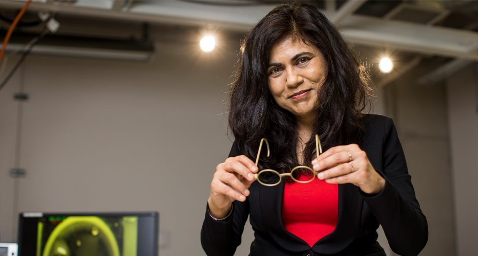 An olive-skinned woman in a red top and black blazer taking off her protective eyewear. 